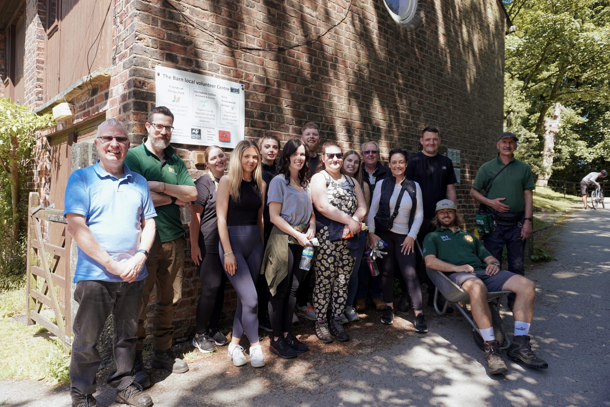 A group of people pose for a photo outside a brick building near a sign and some trees on a sunny day.
