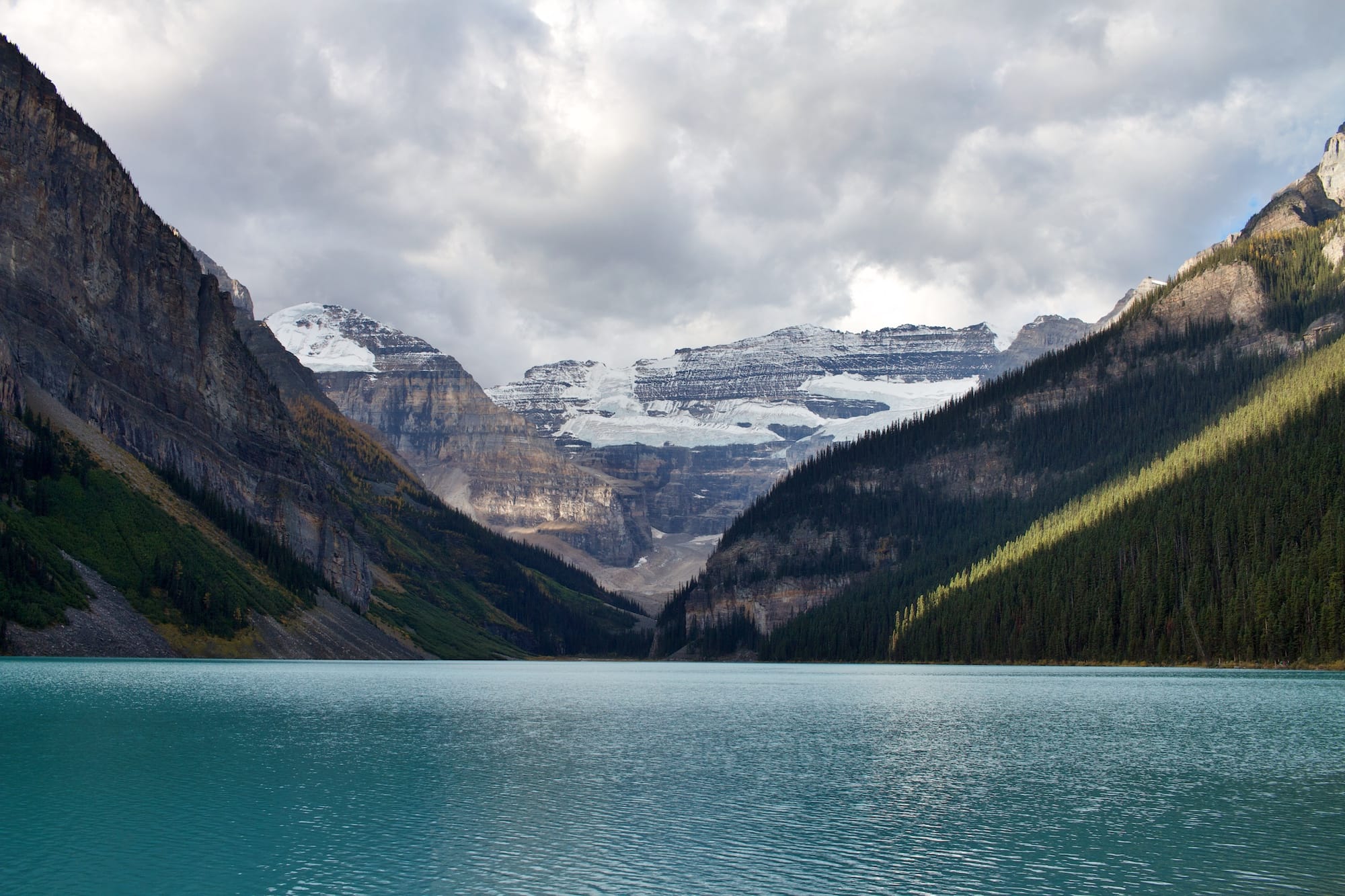 A turquoise lake, surrounded by forested mountains with snow-capped peaks under a cloudy sky, offers inspiration for environmental consultancy focused on preserving such pristine natural beauty.
